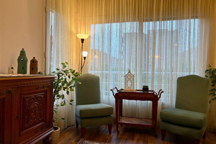 Two green armchairs, a carved wooden cabinet and a lantern beside tall curtained windows